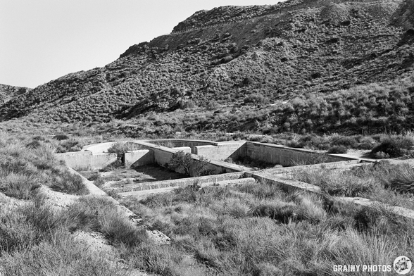 A desolate landscape featuring the remains of concrete structures set against rocky hills, surrounded by sparse vegetation in black and white. The scene evokes a sense of abandonment and the passage of time.