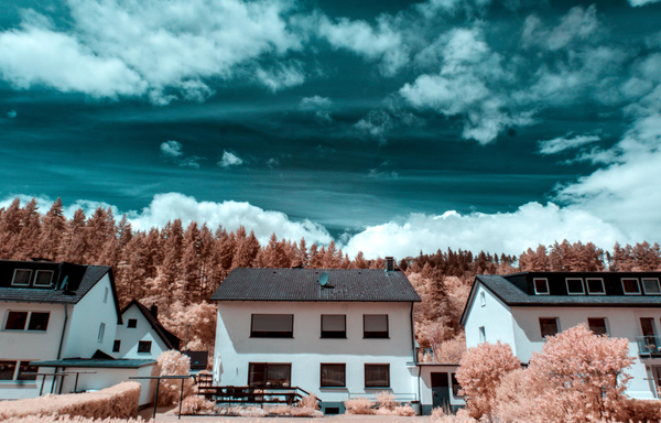 Infrared landscape with three white houses surrounded by orange foliage, with a teal-blue sky and scattered clouds above them.