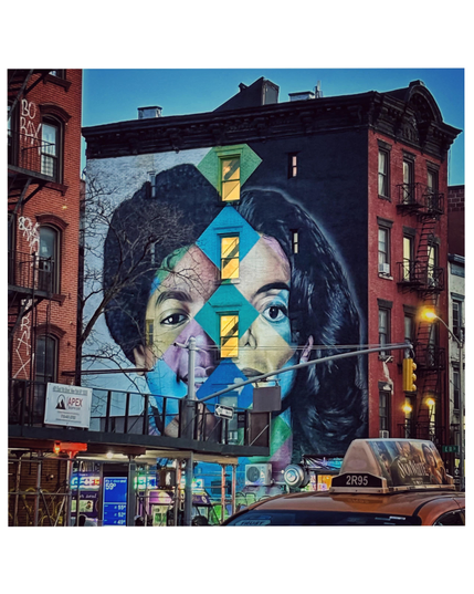 A large mural of Michael Jackson on the side of a five-story building in Manhattan’s East Village (First Avenue and 11th Street) by Brazilian artist Eduardo Kobra. The artwork features a giant split face with half of MJ’s boyhood face against a light background on the left, and half of his post-plastic-surgery adult face against a dark background on the right. Dividing the two faces is a vertical row of windows surrounded by a multicolored diamond pattern and showing the silhouettes of staircases inside. Around the building are the usual city elements: streetlights, scaffolding, the top of a taxicab, etc.