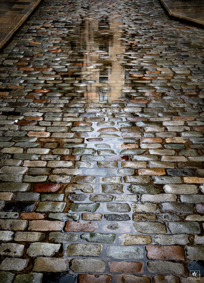 Color photo of a rain puddle in a colorful cobblestone street reflecting the facade of a yellow building.