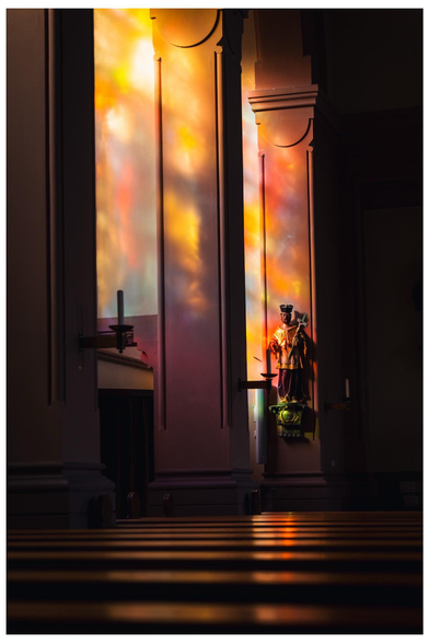 A soft light filters through stained glass, casting colorful reflections on the church wall, where a statue is prominently displayed. Wooden pews are visible in the foreground, creating a serene atmosphere.