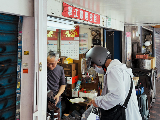 一名戴着头盔和口罩的送货员在店铺前与店主交流，店铺里堆满了箱子和商品。