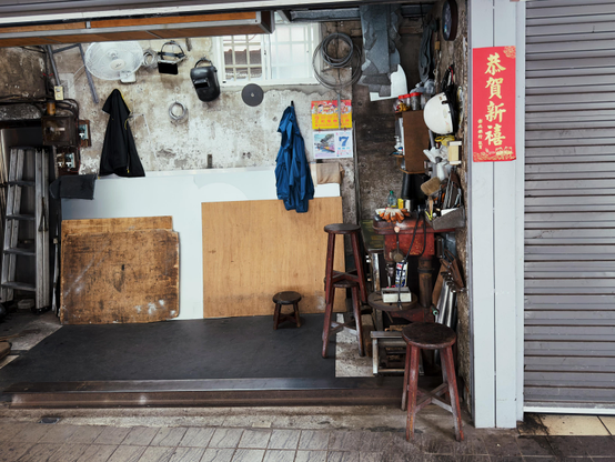 这个店铺内部很杂乱，有各种工具、木板和椅子，门口挂着红色的祝贺新年的横幅。