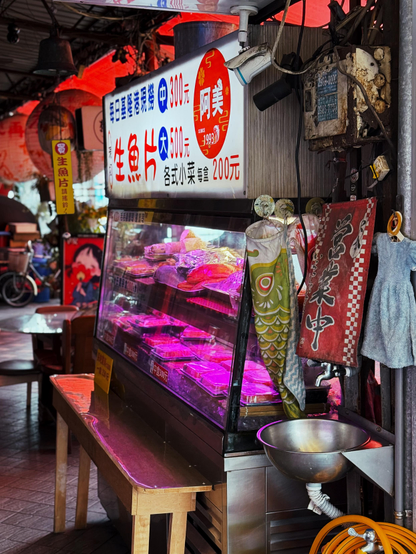 展示一個街邊店鋪，裡面有冷藏櫃內放著生魚片，店面標誌和布條上寫有價格和食品信息。
