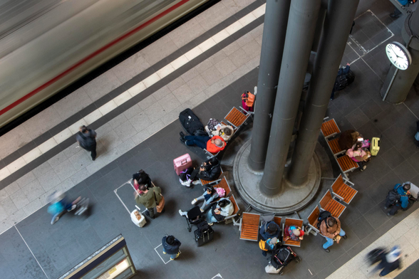 Photo of a train platform seen from above, with seats arranged around tall pillars, with people sitting in the chairs and milling around them. In the top left a train passes. The photo is taken with long exposure so the moving train and the people moving are blurred.