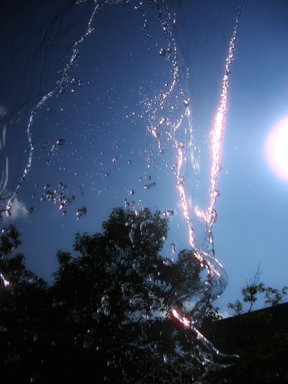 Looking through the water cascading down from a street corner water feature. The clear blue sky and a hazy distorted tree can be seen as well as the sun in the top right edge.
