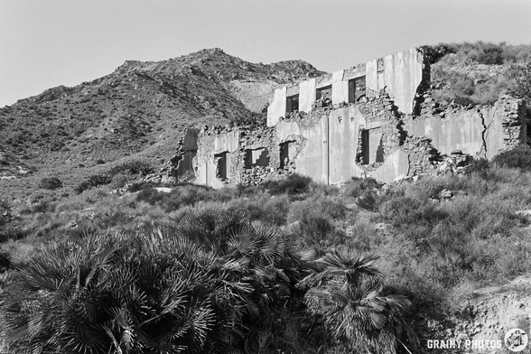 A weathered, abandoned structure stands on a hillside, surrounded by lush greenery and rugged mountains. The building's crumbling walls and open windows hint at its long-forgotten history amidst a desolate landscape.
