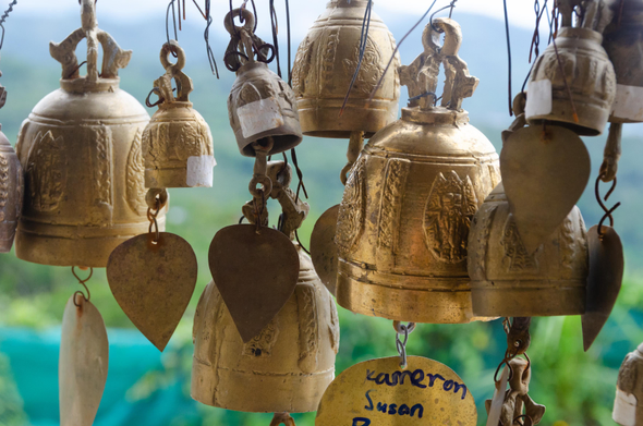 a close-up photo of temple bells in a Buddhist temple in Thailand, somewhere around Phuket. the crowded bells are ornate with Buddhist imagery. one of the tags carry the names "Kameron and Susan". lush green and blue landscape can be seen blurrily in the background