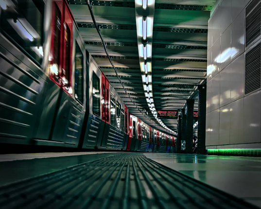 Links im Bild steht ein Zug der Hamburger U-Bahn, nur die Seite mit den charakteristischen roten Türen ist zu sehen. Von vorn zieht sich der Bahnsteig in einem rechtsgeneigten Bogen in den Bildhintergrund. An der Decke folgen die kalten Neonlichter dem Bogen. Rechts an der Wand ist in Bodennähe ein grüner Leuchtstreifen zu sehen, der wohl auch nur dekorativen Zwecken dient. Kein Mensch ist auf dem Bahnsteig zu erkennen... 
