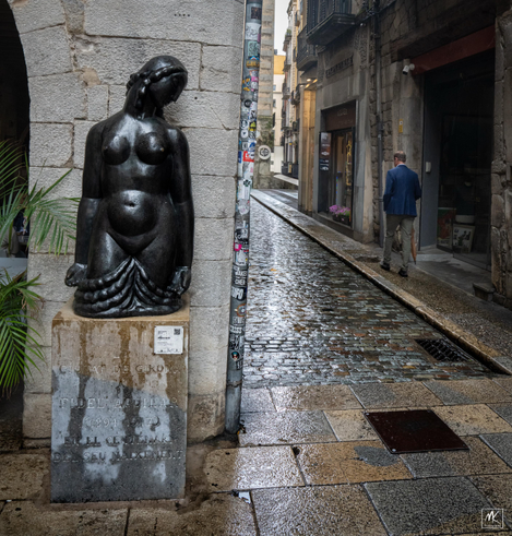 Color photo of a black bronze statue (copy of the original by Fidel Aguilar titled “Imploració”) on a street corner that is of female figure from the lower thighs up mounted on a rectangular granite block.  The figure has her head slightly tilted and her torso naked. Her face is surrounded by a braid that falls down her neck and is presented closed eyes and in profile facing towards the corner.  On the street beyond in the middle distance a well dressed man carrying an umbrella is walking away. 