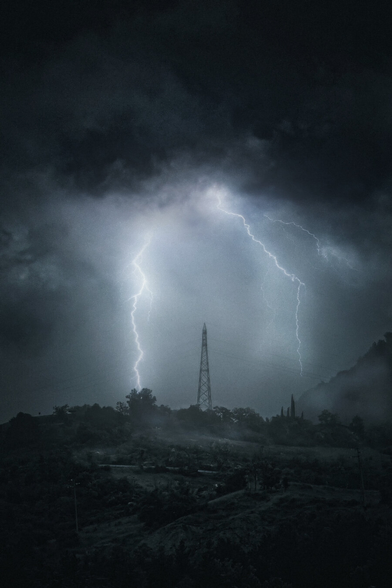 This is a photograph of a dramatic, stormy night scene. A tall metal electricity tower stands in the middle against a dark, cloudy sky. It is illuminated by two bright, jagged lightning bolts. The foreground is misty and foggy landscape with scattered trees. The photo is dark with a blue-grey color palette.