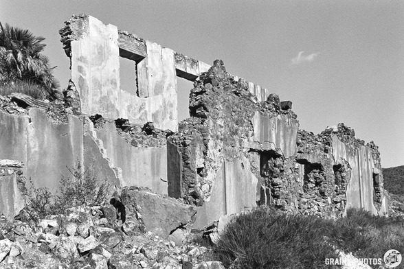 Black and white photograph of dilapidated ruins with crumbling walls and openings. Surrounding vegetation includes rocks and sparse greenery, highlighting the decay of this historic structure set against a clear sky.