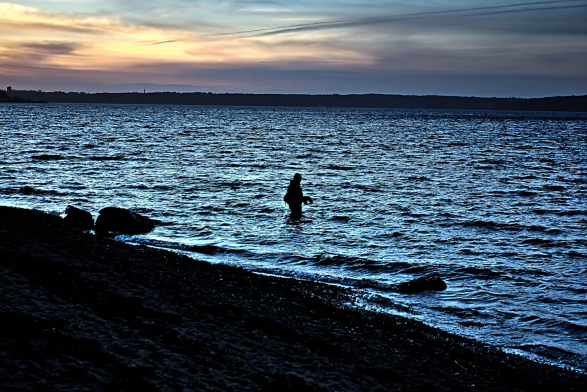 Am Ufer des Meeres steht ein Fischer einige Schritte tief im Wasser. Der Himmel zeigt sich im Sonnenuntergang bedeckt und schenkt uns ein Farbenspiel aus rotgoldenen, blauen und dunklen Tönen. 