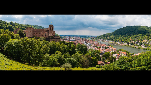 Blick über eine grüne Landschaft. Links auf einem Berg ein teilweise verfallenes Schloss, weiter Rechts eine Stadt an einem Fluss. Im Hintergrund dramatische Wolken.