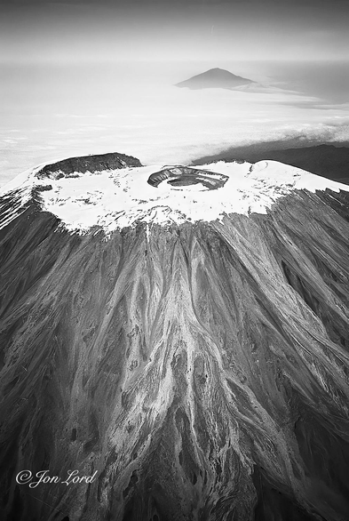 This is a black and white aerial photo in portrait format of the snow covered summit of the dormant volcano Mount Kilimanjaro (5895m) and looking to the west with a further dormant volcano, Mount Meru (4562m) in the background. Tanzania (1988). 

In the foreground is the African volcano's summit, viewed from the east with only the upper portion in view and dominating the foreground of the image. It's steep almost conical sides are heavily eroded with time and heavy tropical rains. The surface being made up of multiple deep grooves and with the appearance of numerous earth and rock avalanches running down the mountain. At first look, the snow covered summit looks perfectly flat with a round caldera in the centre, though there might be multiple calderas, one within another and so on. Beyond Kilimanjaro is a layer of flat, brilliant white stratus cloud obscuring the ground. In the distance and rising up through the clouds, 70km away, is a further dormant volcano: Mount Meru. Again it has a conical shape to it but more rounded or flattened. Beyond is a horizon with a milky white sky above becoming darker with increasing height. 