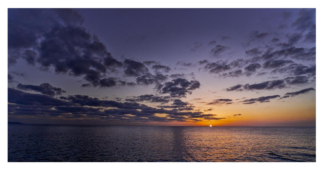 This is a tranquil and atmospheric seascape photograph taken at sunset. The lower half of the image shows a wide, calm ocean that stretches all the way to the horizon. The water is dark blue with gentle ripples and catches a warm, glowing reflection of the setting sun — a bright orange-gold path of light that leads from the horizon directly toward the viewer across the surface of the sea. On the horizon line, slightly to the right of center, sits a small, bright white-orange sun just above the water. Around the sun, the sky glows with warm colors: vivid orange and golden yellow that gradually fade upward into soft pink, lavender, and finally into deep indigo and dark blue at the top of the image. The sky is filled with dramatic, layered clouds. Some clouds are dark and heavy, especially in the upper part of the frame, creating strong contrast. Others are illuminated from below by the sunset, showing softer edges with shades of purple, gray, and orange. The clouds give the sky a rich, textured, and moody feeling. Overall, the photograph has a peaceful yet dramatic mood — the vastness of the ocean meeting a colorful, cloudy sky at the moment of sunset.