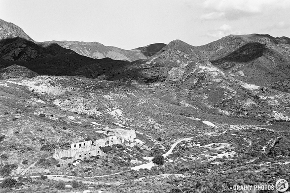 A desolate landscape featuring rugged mountains and a dilapidated building nestled in a valley. The image showcases a monochromatic view highlighting the texture of the terrain and the remnants of human habitation amidst nature.