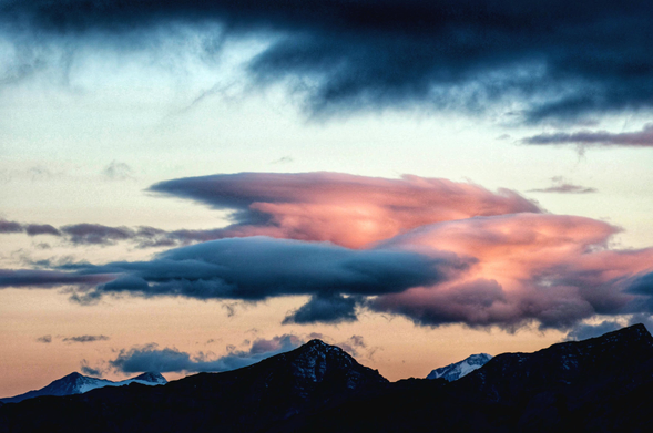 Diese stimmungsvolle Landschaftsaufnahme zeigt ein weites Gebirgspanorama während der Dämmerung unter einem dramatisch bewölkten Himmel. Im unteren Bilddrittel erstreckt sich die dunkle, scharfkantige Silhouette einer Bergkette, deren massivster Gipfel sich in der Mitte emporhebt und an den Flanken leichte Schneereste aufweist. Über den Bergen entfaltet sich ein faszinierendes Wolkenspiel in verschiedenen Schichten. Das zentrale Element ist eine große, horizontal gestreckte Wolkenformation in der Bildmitte, die durch das letzte Licht der untergegangenen Sonne in sanften Rosa- und Pfirsichtönen leuchtet und an der Unterseite von dunkleren, graublauen Schatten akzentuiert wird. Diese leuchtende Wolke wirkt fast wie ein schwebendes Plateau. Darüber und an den Rändern dominieren schwere, tiefblaue und fast schwarze Wolkenmassen, die einen starken Kontrast zum helleren, blassgelben bis zartblauen Abendhimmel im Hintergrund bilden. Die gesamte Szenerie strahlt eine kühle, aber durch die Farbtupfer auch friedliche und majestätische Atmosphäre aus.