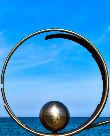 A large metal sculpture with a steel ball sitting at the inner bottom of a larger ring stands in front of a blue sky and the Mediterranean Sea.
