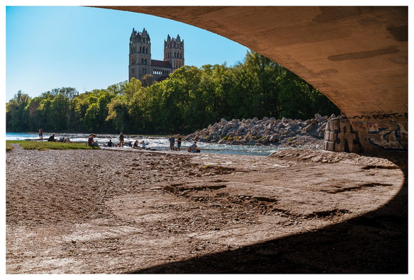 Wir stehen unter einer Brücke die über die Isar führt. Ihre Rundbögen werfen einen bogenförmigen Schatten auf den Boden. Im Hintergrund der Isarstrand, Personen am/im Wasser, Bäume und eine neoromanische Kirche mit Doppeltürmen.