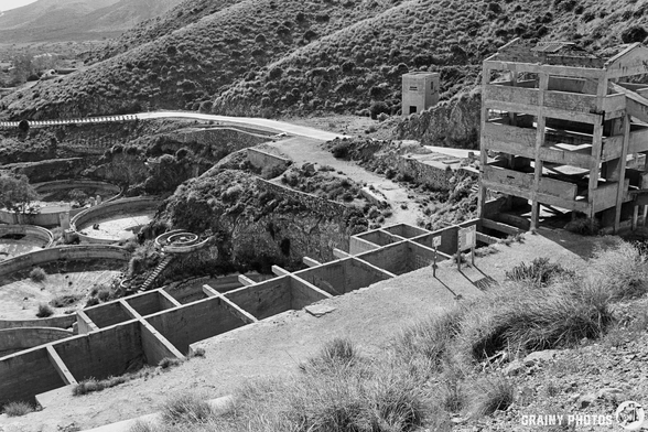 A black and white image of an abandoned landscape featuring dilapidated buildings, overgrown vegetation, and a winding road cutting through a hilly terrain.