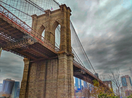 A view looking up at the underside of the majestic Brooklyn Bridge. The stone arches dominate the left side of the photo, with its suspension cables cutting across a moody, overcast sky. The Manhattan skyline and trees are visible at the bottom.