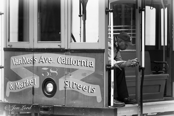 This is a black and white candid street photo in landscape format of a cable car operator sitting on a bench at the front of the stationary car, leaning forward and looking at his mobile (cell) phone. San Francisco, California. (2015).

The cable car is viewed obliquely with the front (or could be rear?) and part of the side in view, the top and base are out of sight. The left half of the photo is filled with the front (or rear) of the cable car. There are three vertical rectangular windows for the 'Grip' (driver) to see from. Below are three wood panels with a painted banner reading 'Van Ness Ave.. California & Market Streets' and with a single headlight and the number '58' above the light. Sitting on a dark wooden bench that stretches along the side of the car is our 'Grip' or possibly the conductor on his break. He is an African-American male, middle-aged, wearing a baseball cap, light coloured jacket, dark trousers and heavy boots. He is leaning forward and looking at his mobile that he's holding in his left hand and is holding his right hand above the phone as if about to scroll or type. To his left is a long, vertical white pole for standing passengers to hold on to. 

The location: California Street, San Francisco.