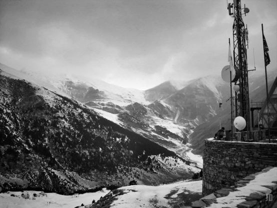 La imagen muestra un paisaje montañoso nevado en un día nublado. A la derecha hay una estructura de piedra con antenas y banderas, mientras que al fondo se extienden montañas cubiertas parcialmente de nieve y bosques en las laderas. La escena transmite una sensación de frío y tranquilidad.