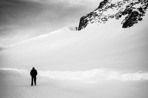 A lone figure walks on a snowy landscape, with a large snow-covered mountain in the background. The image is in black and white, highlighting the stark contrast between the snow and the rocky terrain. The scene conveys a sense of solitude and vastness.