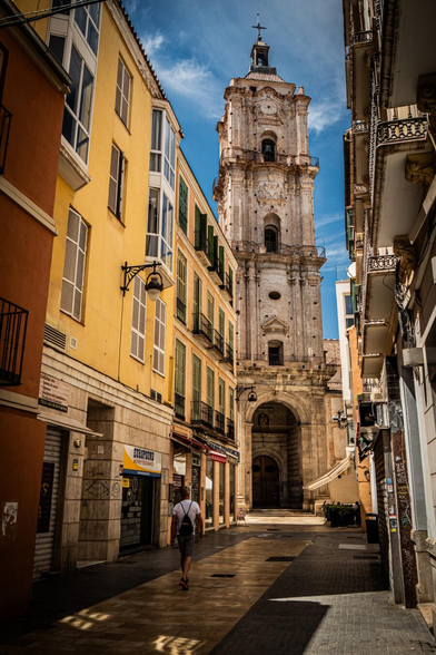 A narrow paved street in Málaga lined with vibrant yellow and orange buildings leads to toward the tall baroque bell tower of San Juan Church rising into a blue sky.