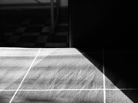 A black and white photo of a tiled table with sunlight casting shadows on it. The background shows a checkered floor and some furniture.