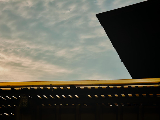 Cloudy sky with the silhouette of a building's roof and a gutter in the foreground.