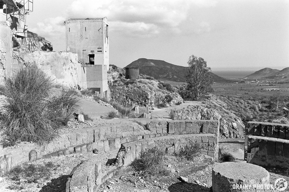 A black-and-white photograph of a desolate landscape featuring abandoned structures, part of the abandoned Rodalquilar Gold Mines, rocky terrain, and distant hills leading to the ocean. The scene conveys a sense of decay and historical significance amidst nature's encroachment.