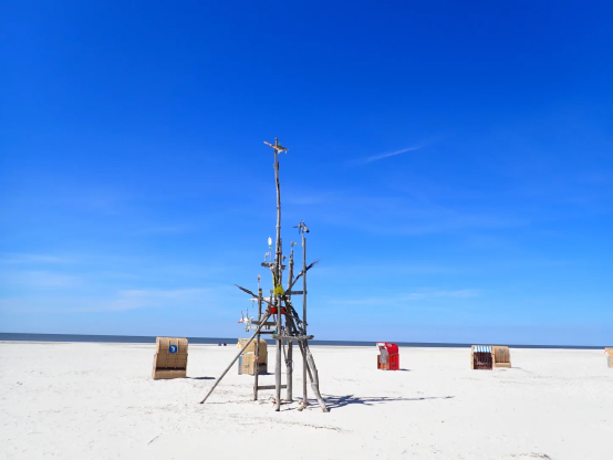 heller Sandstrand unter einem blauen Himmel. Im Hintergrund die Nordsee. Auf dem Strand stehen drei Strandkörbe und eine Installation, die anscheinend aus Treibgut zusammengebaut wurde.