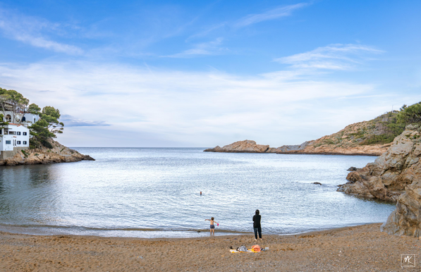 Color photo of a cala (Catalan for cove) with a sandy, pebbly beach. On the left side are some white washed houses above a rocky shore and on the right side are rocky cliffs and slopes. On the beach is a woman and a child standing looking out at the water where the head of a swimming man is sticking up above the surface.