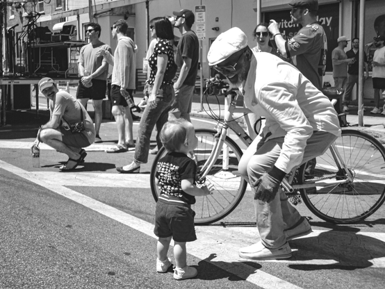 A smiling man standing beside a bicycle dances and smiles toward a baby at the Italian Market Festival in Philadelphia.