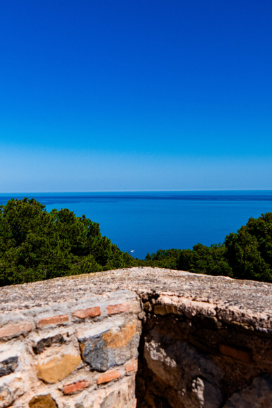 A scenic view of the blue Mediterranean Sea under a blue sky. The two blues meet at the horizon while in the foreground leafy green trees can be seen behind a stone wall corner at Castillo de Gibralfaro in Málaga, Spain. The corner creates an arrow of sorts pointing toward the horizon.