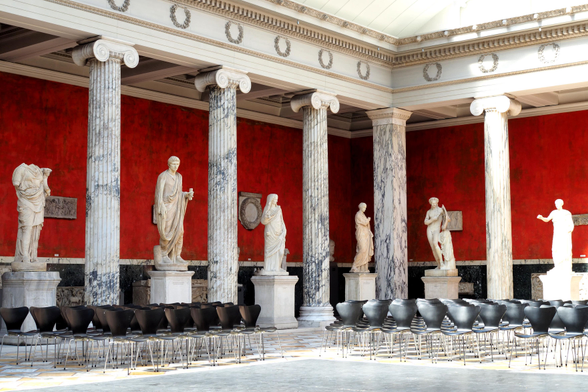 Lebensgroße weiße Stein-Skulpturen in der Glyptotek in Kopenhagen stehen zwischen weißgrauen Säulen vor roten Wänden. Im Raum davor viele leere Stühle mit schwarzen Sitzflächen. Der Boden und die Decke sind hell.

Life-size white stone sculptures in the Glyptotek in Copenhagen stand between white-grey columns against red walls. In the space in front of them are many empty chairs with black seats. The floor and ceiling are light in colour.