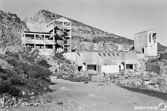 A desolate, abandoned concrete structure, part of the abandoned Rodalquilar Gold Mines, sits on rocky terrain, surrounded by mountains. The building shows signs of decay, with broken walls and a rugged landscape, reflecting a sense of neglect and the passage of time.