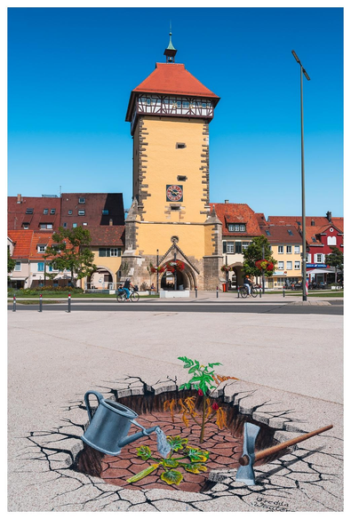 Ein mit Kreide auf Asphalt gemaltes Bild, welches ein Loch im Asphalt darstellt. Aus dem Loch wachsen Pflanzen. Aus einer gemalten Gieskanne fließt wasser auf eine Pflanze. Eine gemalte Hacke liegt neben dem Loch. Im Hintergrund einige ältere Häuser und ein hoher Wehrturm.
