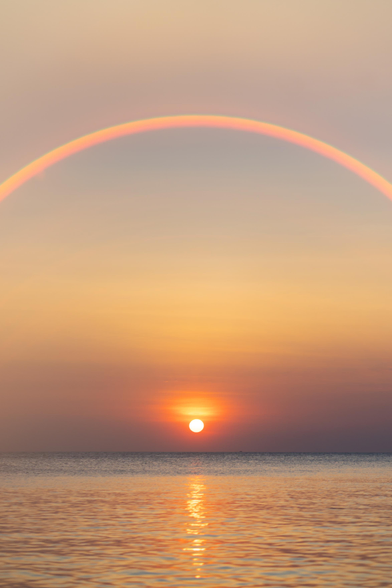 I took this photo at sunset on Ko Samet in Thailand. we were lounging on a jetty and I had my lens filter on. the photo shows the ocean with the setting sun just above the water, casting a line of bright reflection in the water. the lens filter results in a round arc lens flare above the sun
