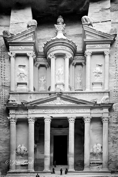 This is black and white photo in portrait format of the ancient Al-Khazneh also know as the "Treasury" and sometimes the Khaznat el-Far'oun, a vast, sandstone, rock-cut tomb in the ancient Nabatean city of Petra in Jordan (1996).

The photo is taken looking straight at the facade, from a distance of about twenty or thirty metres, The structure fills almost all of the image. The opening to the Al-Khazneh or "Treasury" is framed by six tall columns three either side. Above the columns is a lintel with erroded carvings stretching the width of the columns and structure. Between and above the inner four columns is a pitched roof, extending outwards, perhaps a few metres again with numerous eroded carvings on the surface. Above is a further and much thicker lintel supporting six further columns carved and arranged in pairs. The outer pairs support a triangular, roof like structure with a worn carvings between the columns. The middle two columns make up a cylindrical centre piece topped with a crown-like carving with a peak in the middle. Between and to the sides of the centre two columns are statue like reliefs of people. Above is a void before the sandstone cliff continues upwards. At the base are five people, tourists, guides or traders? with a dark vertical rectangular opening to the tomb. 

Tourism has taken its toll as the Khazneh surface has receded by 40 mm in less than ten years from touching, leaning, or rubbing on the soft sandstone walls.