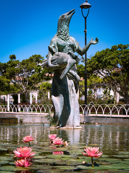 A large bronze pond statue featuring a figure with a woman's body and a bird's head looking upward. She holds a large water bird in her arms. In the foreground, vibrant pink water lilies float on the pond's surface with green trees in the background.