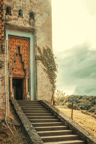 A photograph of an ancient looking stone building with a weathered facade (it was actually only built in the 19th century!). There is a carved door frame and mantle with intricate geometric patterns in orange. A worn stone staircase leads up to the door. There are two small stone.lion statues stand on either side of the doorframe. The background has a cloudy sky and a distant, hilly landscape. The image has a warm tone. (Was trying to bring out the dusty/desert/vintage vibe, you know?)
