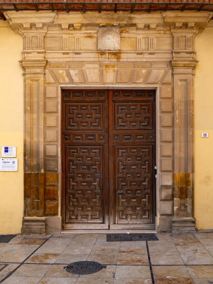 A symmetrical shot of the entrance to Palacio de Villalcázar. It features a heavy, dark brown double-leaf wooden entrance door with intricate square paneling. The door is framed by a light-colored, rustic stone archway and a pale yellow wall. Plaques are visible on the left wall, and the number 23 is on the right.