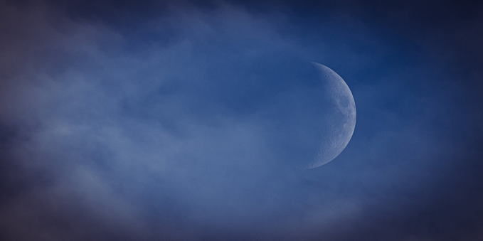 A wide aspect ratio photograph of the moon, slightly less than half. The moon is placed quite far to the right in the photograph and the illuminated side is towards the right as well. The surrounding sky is deep blue and the entire photo is covered by a light cloud cover, thickest toward the left, balancing the moon to the right.