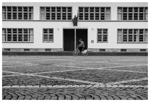 Blick über einen leeren Platz. Vor einem Gebäude mit auffällig kleinkarierter Fensterfront fährt ein Mann auf einem Leihfahrrad. Am Gebäude steht "DEM LEBENDIGEN GEIST".