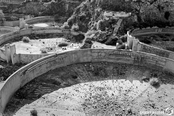 An aerial view of abandoned, circular concrete sediment tank structures surrounded by overgrown vegetation, set against a rocky terrain. The stark black and white imagery highlights the desolation of the area, emphasizing crumbling walls and sparse plant life.