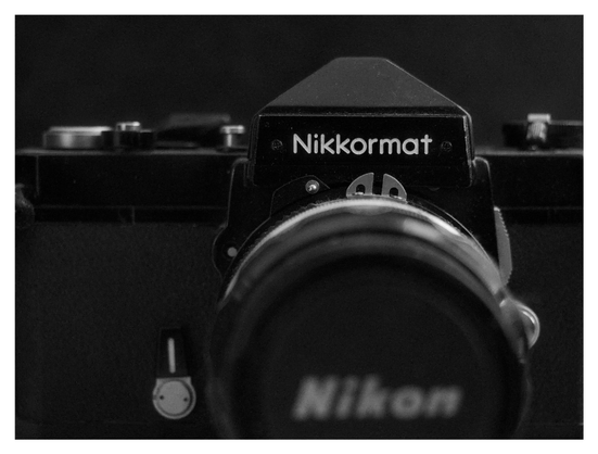 En liggande svartvit bild på en svart SLR-kamera som man behöver film till. På spegelhuset står det "Nikkormat" i vitt och på objektivlocket "Nikon" i vitt.
