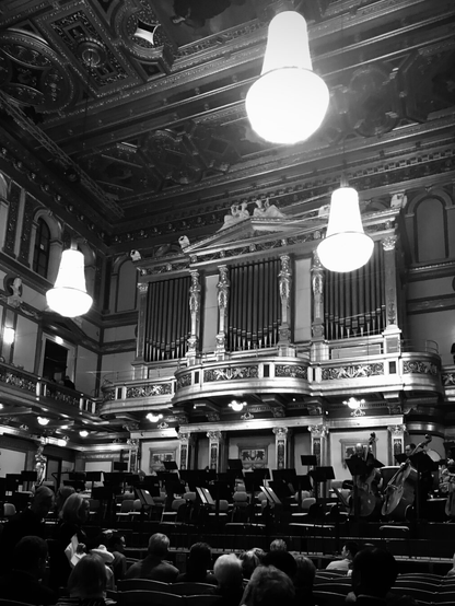 This black-and-white photo shows the large hall of the Vienna Musikverein shortly before a concert. The stage for the musicians is visible, in the foreground rows of seats with guests. In the background you can see the impressive organ. Beautiful crystal chandeliers hang from the ornately decorated ceiling.